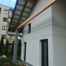 RusztowaniaNDM - Nowoczesny dom jednorodzinny z jasną elewacją, drewnianą konstrukcją zadaszenia i widoczną izolacją przy fundamentach, na tle bloku mieszkalnego.