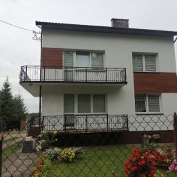 RusztowaniaNDM - Dwupiętrowy dom z białymi ścianami, drewnianymi panelami, balkonem z ozdobną balustradą, widziany zza ogrodzenia z kwiatami.