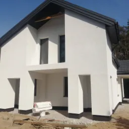 Biały dom z nowoczesną elewacją i ciemną dachówką, widoczne materiały budowlane przed wejściem.