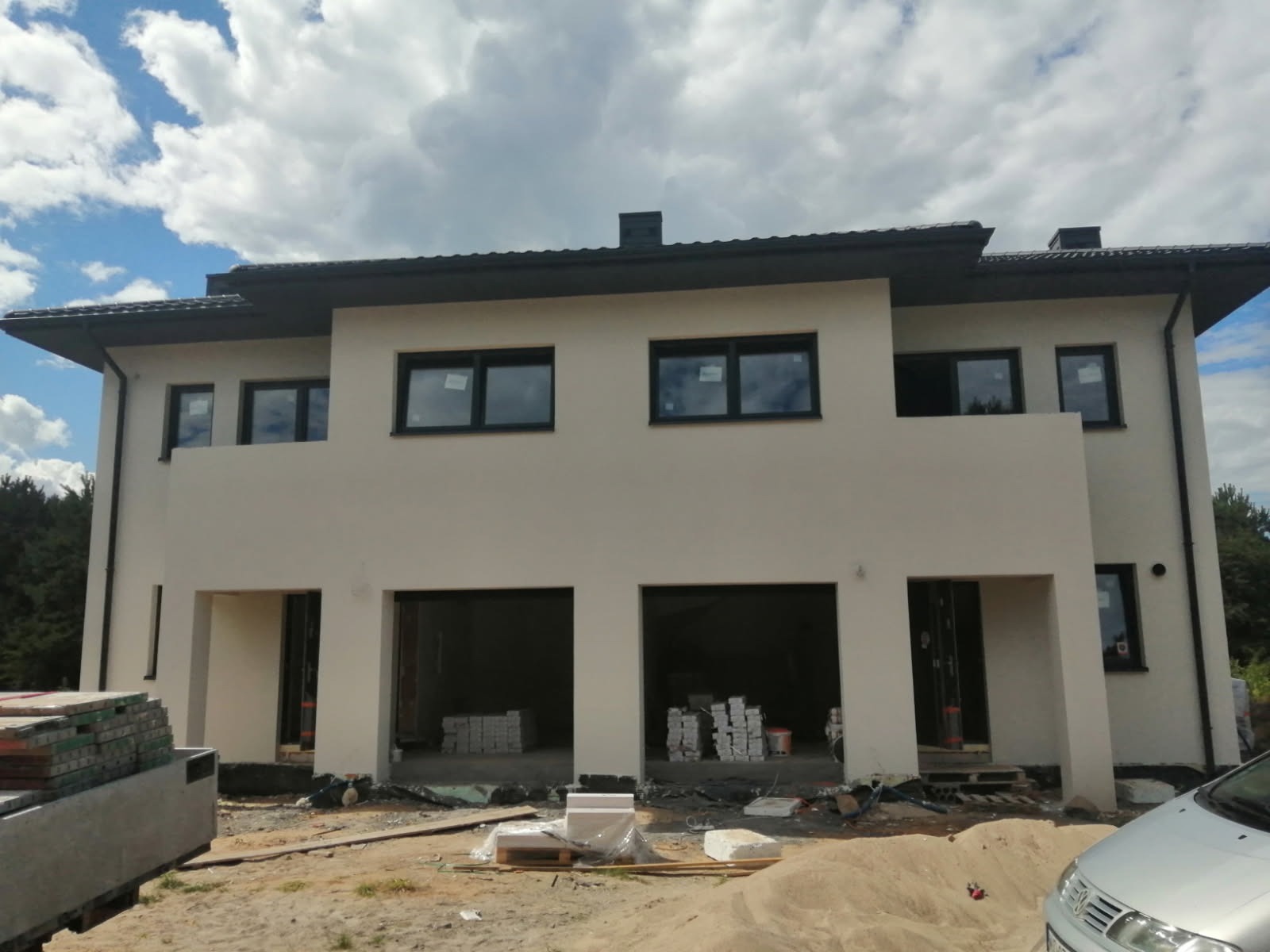 Dwukondygnacyjny dom w trakcie budowy z jasną elewacją, widoczne okna z czarnymi ramami, niezadaszone wnęki, materiały budowlane na placu budowy.