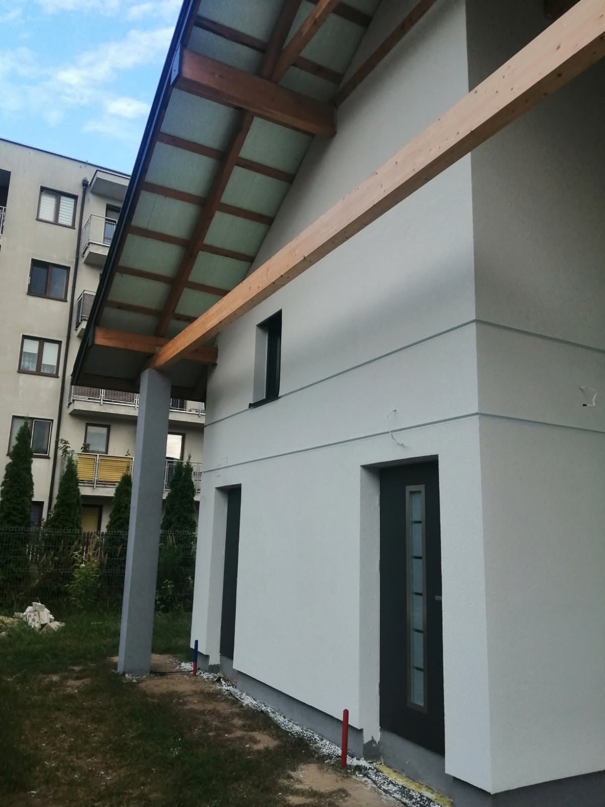 Nowoczesny dom jednorodzinny z jasną elewacją, drewnianą konstrukcją zadaszenia i widoczną izolacją przy fundamentach, na tle bloku mieszkalnego.