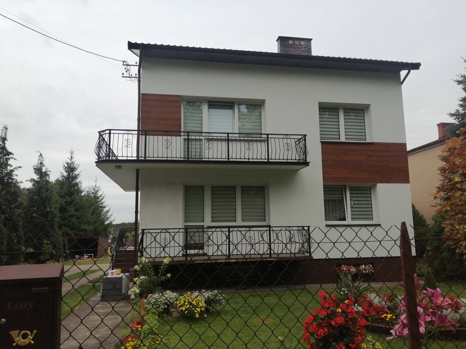 Dwupiętrowy dom z białymi ścianami, drewnianymi panelami, balkonem z ozdobną balustradą, widziany zza ogrodzenia z kwiatami.