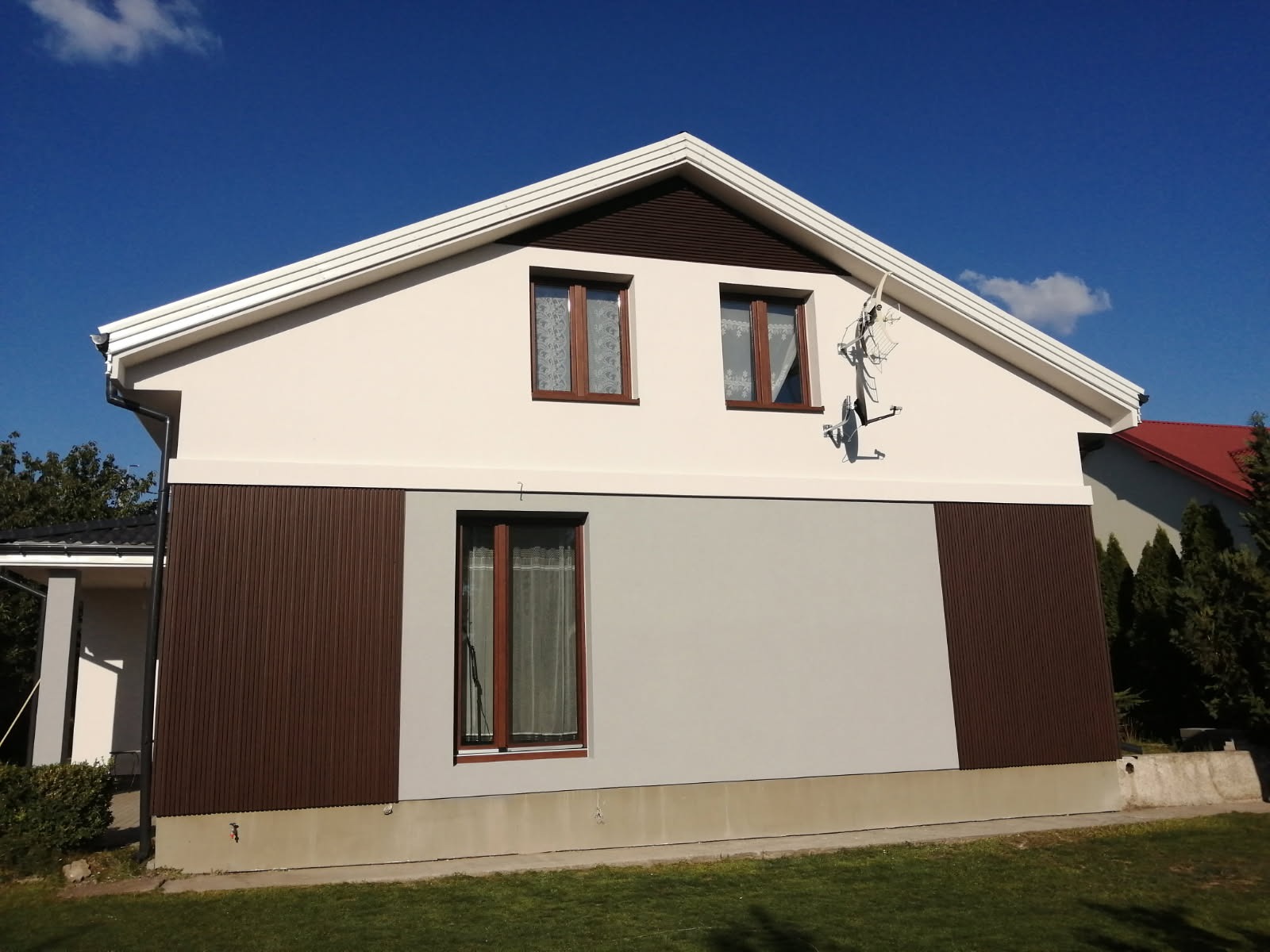 Dom jednorodzinny z nową elewacją: kremowa góra, szara dolna część, brązowe panele boczne, okna z brązowymi ramami i białe obramowanie dachu pod błękitnym niebem.