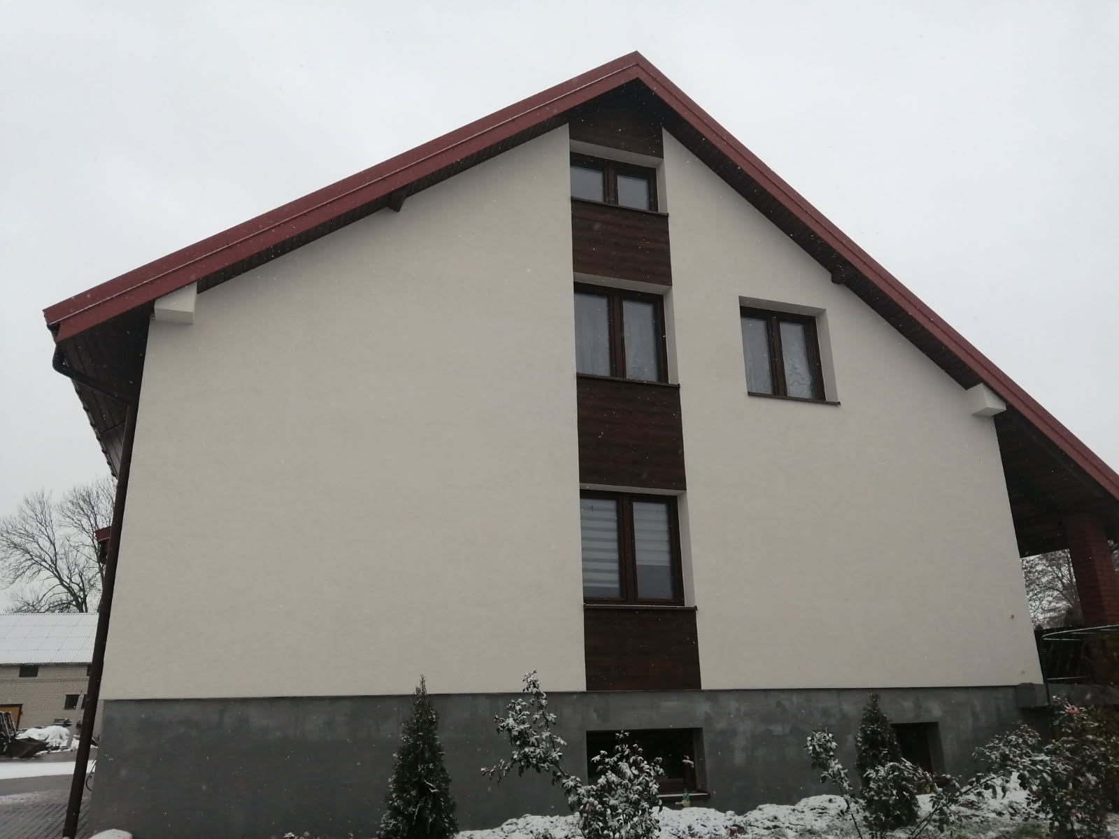 Dwupiętrowy dom z jasną elewacją i ciemnobrązowym dachem w zimowej scenerii, widoczne opady śniegu.