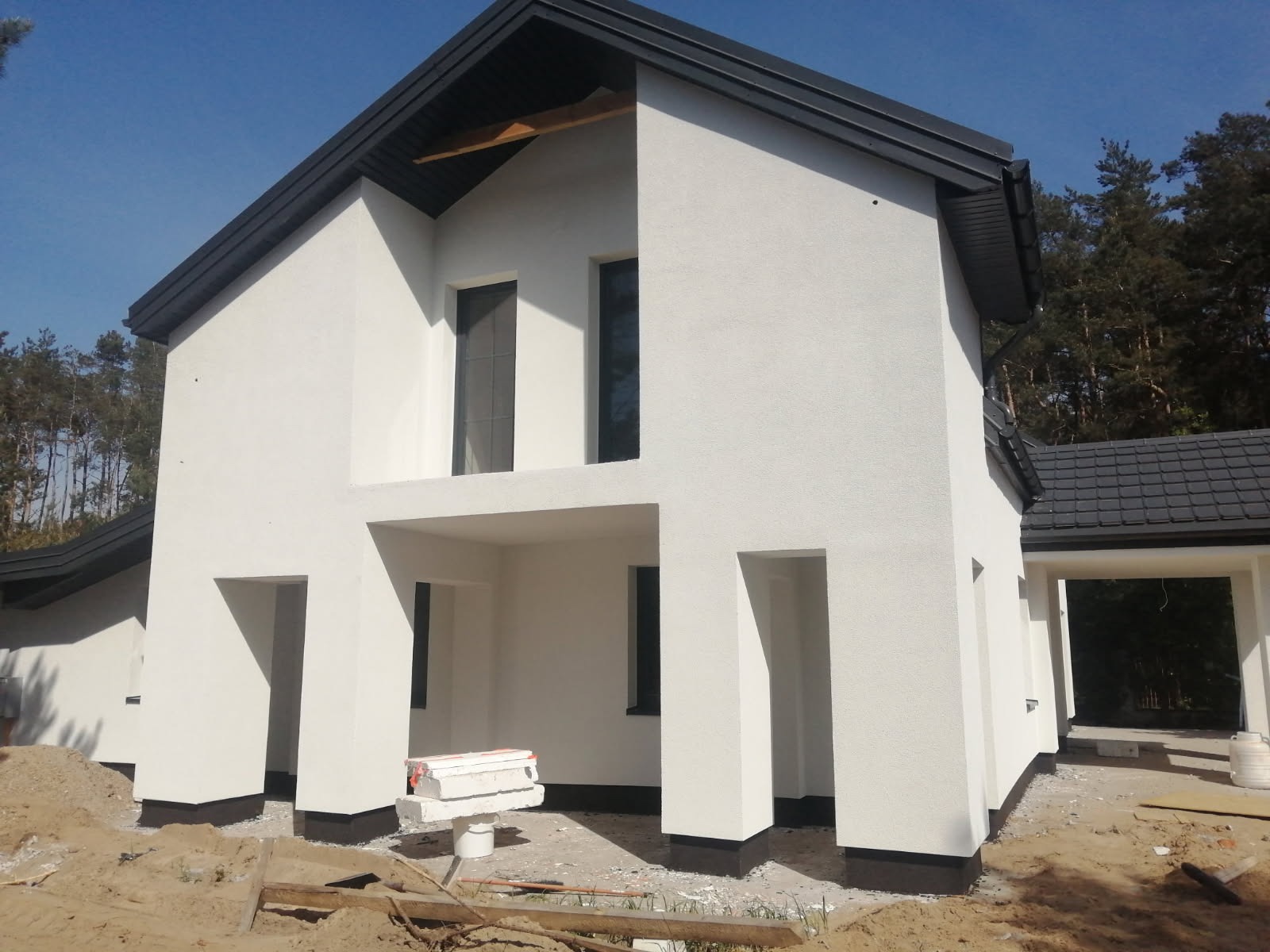 Biały dom z nowoczesną elewacją i ciemną dachówką, widoczne materiały budowlane przed wejściem.
