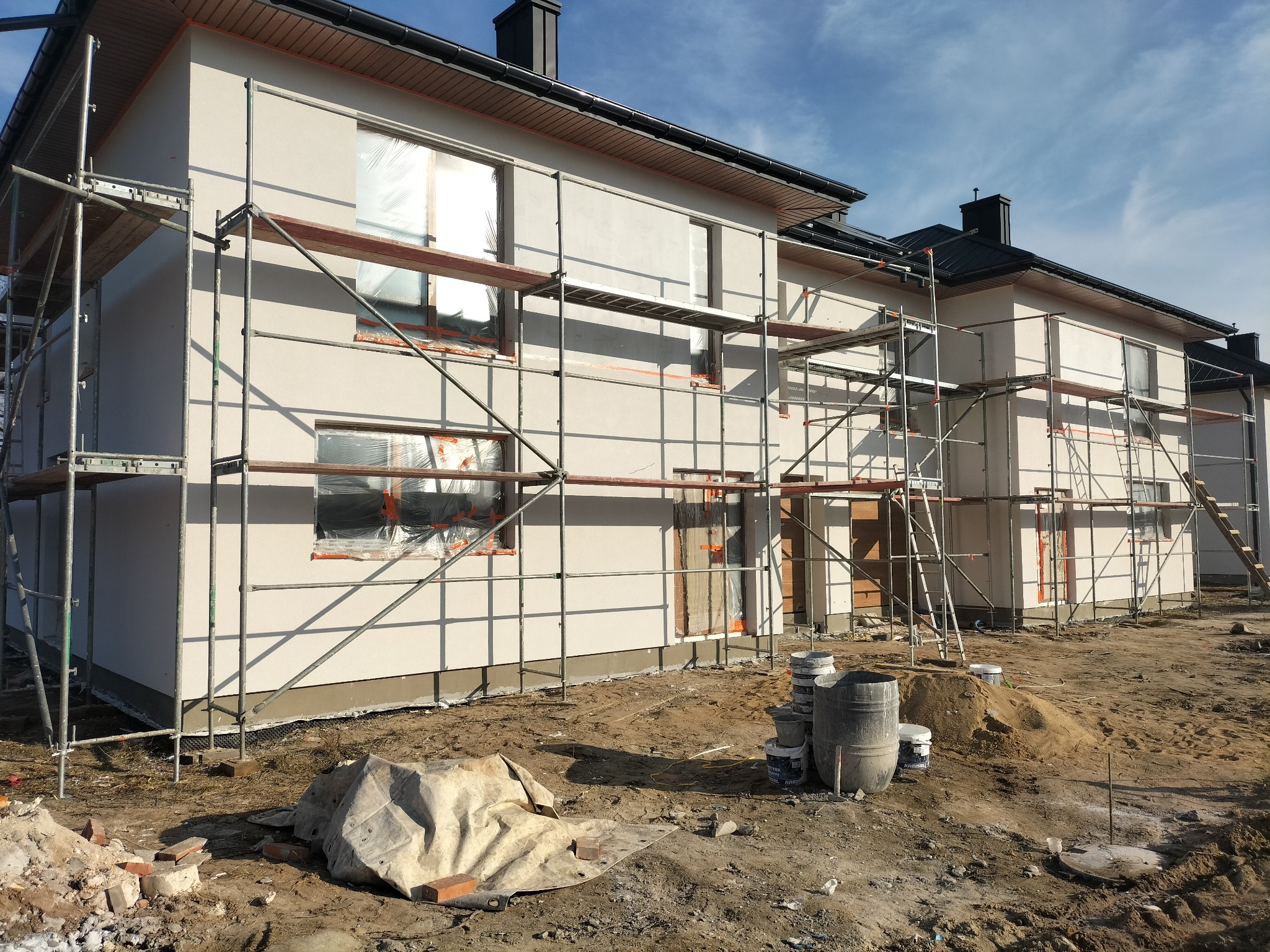 Dwukondygnacyjny dom w trakcie tynkowania elewacji, z widocznymi rusztowaniami, zabezpieczonymi oknami folią i stertą piasku na placu budowy.