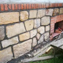 Ściana zewnętrzna budynku wykończona nieregularnymi kamieniami piaskowca z czerwoną cegłą u góry i wnęką, widoczny fragment ławki i gruz budowlany.