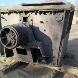 Duża, metalowa konstrukcja przemysłowa z kołem pasowym, umieszczona na drewnianych belkach na piaszczystym podłożu, czyszczona strumieniem ściernym przez osobę w kombinezonie ochronnym.