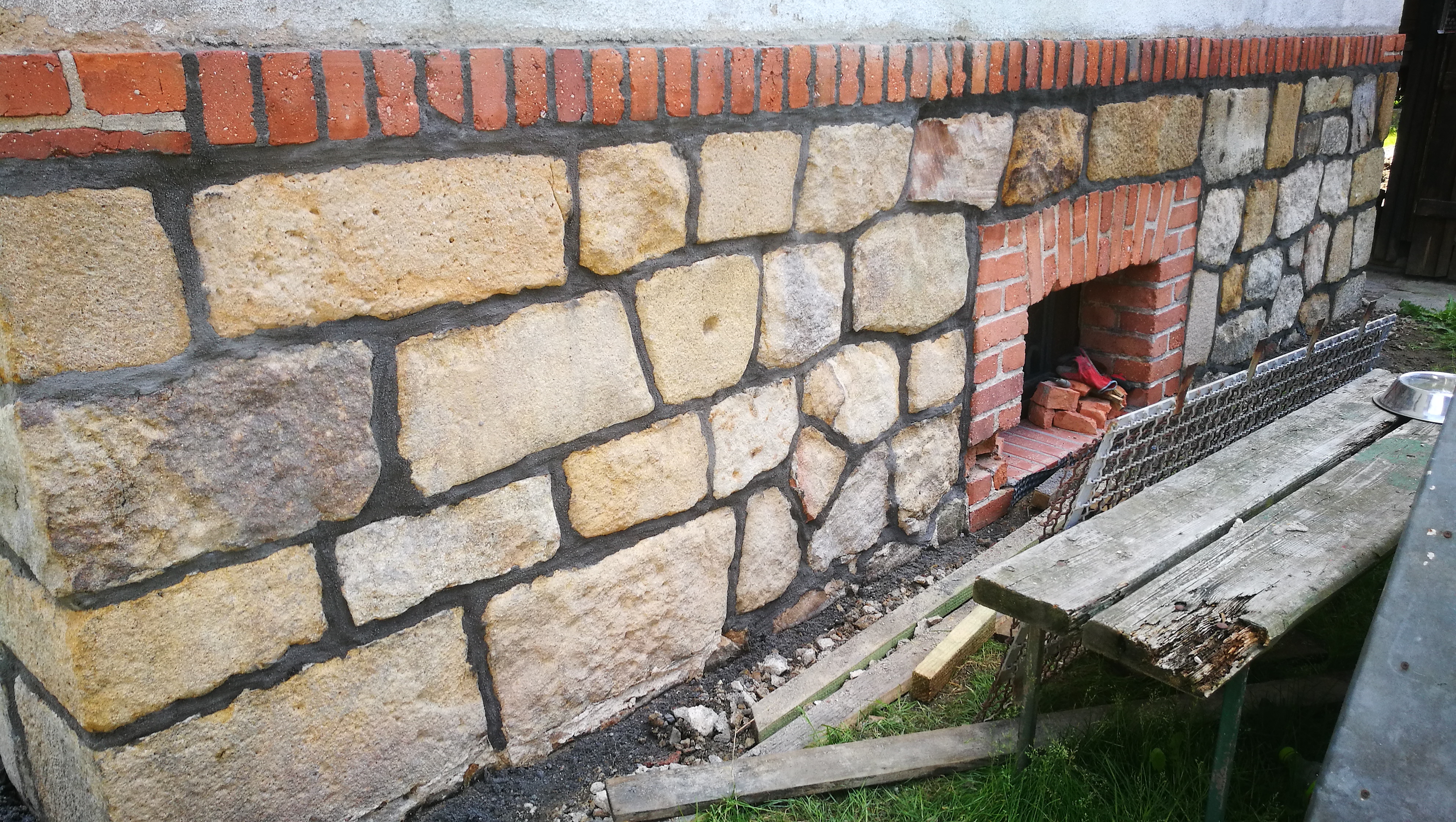 Ściana zewnętrzna budynku wykończona nieregularnymi kamieniami piaskowca z czerwoną cegłą u góry i wnęką, widoczny fragment ławki i gruz budowlany.