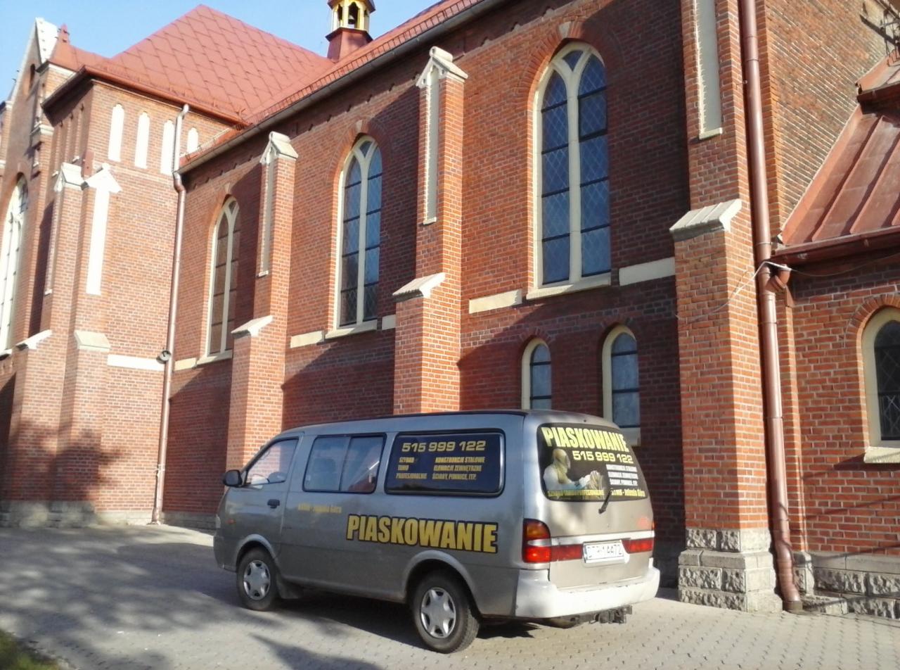 Szara furgonetka z napisem 'Piaskowanie' zaparkowana przed ceglanym kościołem z wysokimi, łukowatymi oknami.