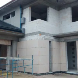 Wznoszony dom z częściowo wykończoną elewacją z jasnych, kwadratowych płyt. Widoczne rusztowanie i surowy stan budynku w tle. W tle niezabudowany teren.