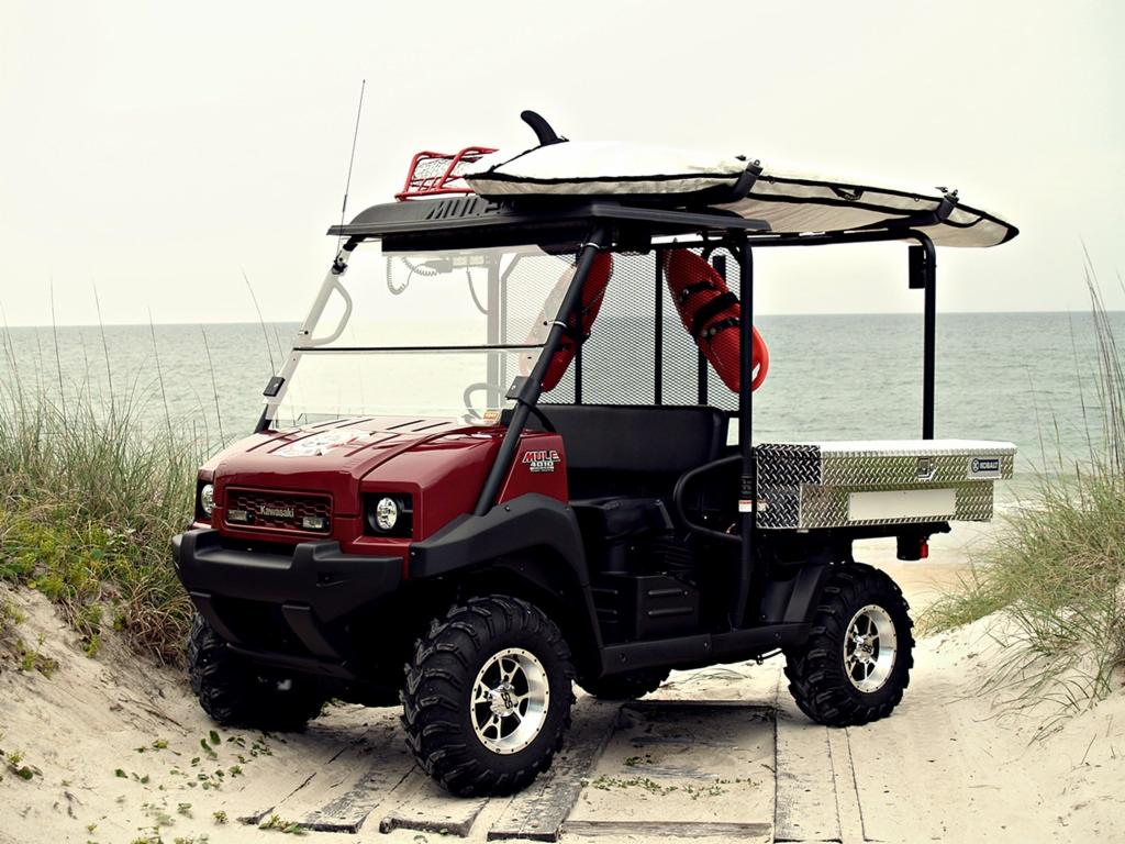 Czerwony pojazd Kawasaki MULE 4010 Diesel 4x4 z deską surfingową i bojkami ratunkowymi na plaży, z widocznym oceanem w tle.