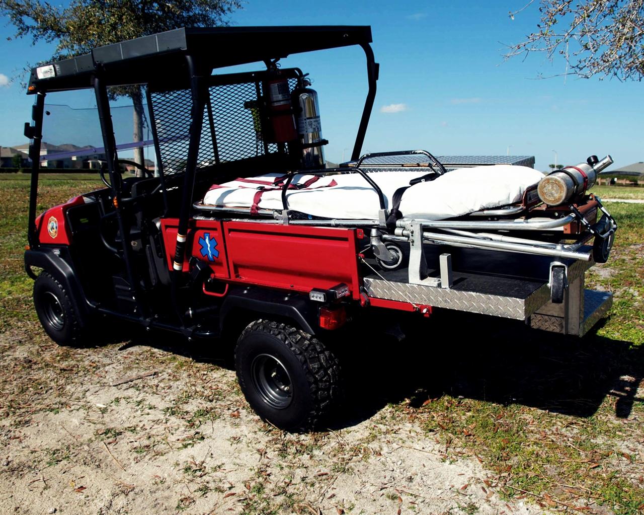 Czerwony pojazd Kawasaki MULE 4010 DIESEL 4X4, zmodyfikowany na potrzeby ratownictwa medycznego, z noszami i butlą tlenową na platformie ładunkowej, zaparkowany na trawiastym terenie.