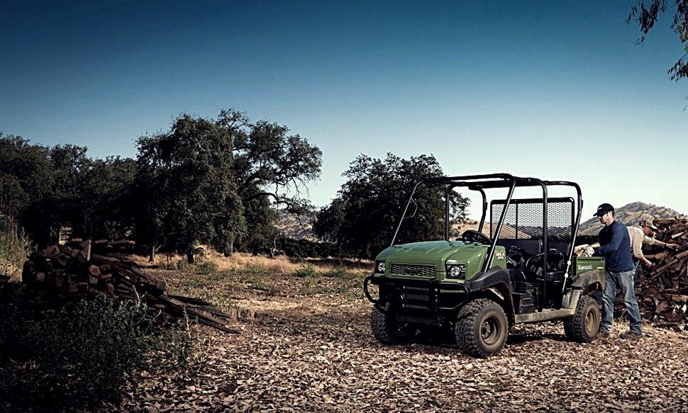 Zielony pojazd Kawasaki MULE 4010 Diesel 4x4 na tle polnym z drzewami, z dwoma osobami w pobliżu sterty drewna.