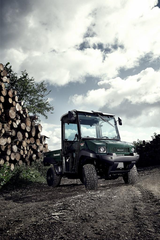 Zielony pojazd Kawasaki MULE 4010 Diesel 4x4 na polnej drodze, z stosem ściętych pni drzew w tle, pod pochmurnym niebem.