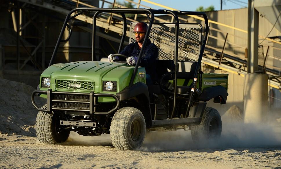 Zielony pojazd Kawasaki MULE 4010 TRANS 4X4 DIESEL z kierowcą w kasku i okularach ochronnych na tle konstrukcji przemysłowej i piasku.