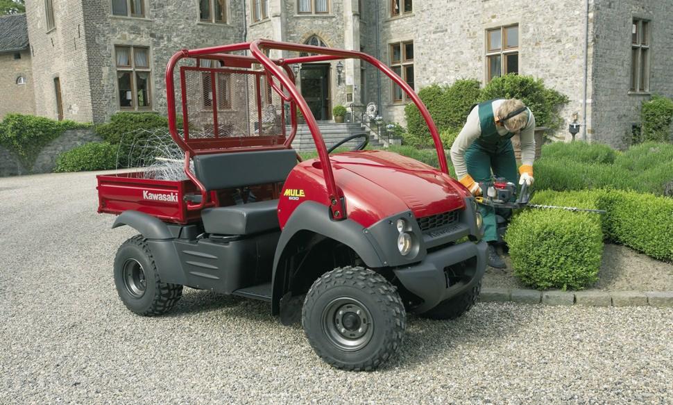Czerwony pojazd Kawasaki MULE 610 4x4 na żwirowym podłożu, obok pracownik w kombinezonie przycina żywopłot za pomocą elektrycznych nożyc, w tle kamienny budynek.