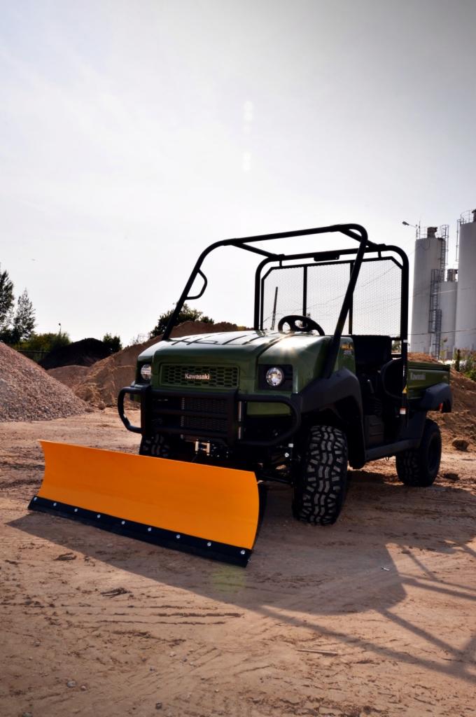 Zielony pojazd użytkowy Kawasaki MULE 4010 Diesel 4x4 z pomarańczowym pługiem śnieżnym, stojący na piaszczystym terenie, w tle hałdy piasku i silosy.