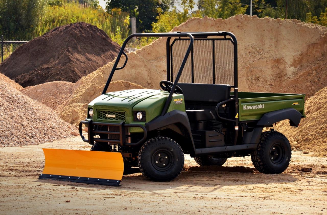 Zielony pojazd UTV Kawasaki MULE 4010 Diesel 4x4 z zamontowanym pomarańczowym pługiem do śniegu, stojący na piaszczystym podłożu z hałdami piasku i żwiru w tle.