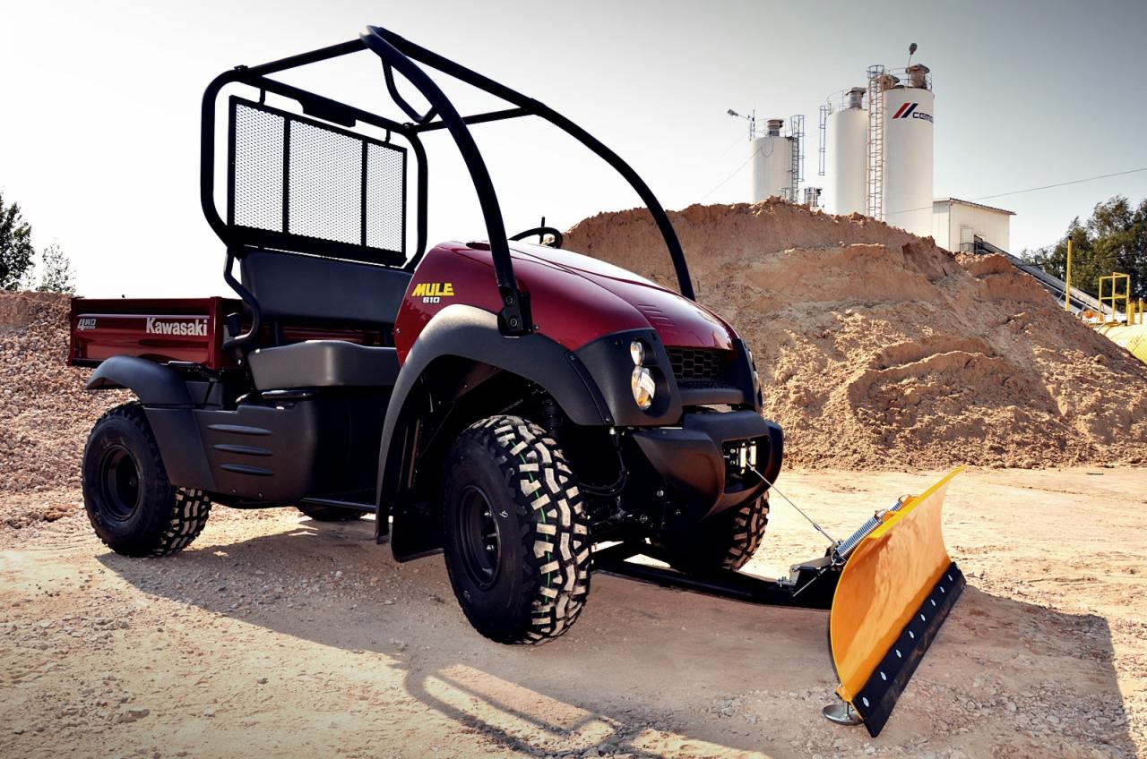 Czerwony pojazd użytkowy Kawasaki MULE 610 4x4 z zamontowanym pługiem śnieżnym na tle hałdy piasku i silosów cementowych.