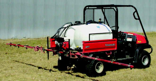 Czerwony pojazd Kawasaki MULE 610 4X4 z białym zbiornikiem i rozpylaczem na polu, widok z boku, z metalową ścianą w tle.