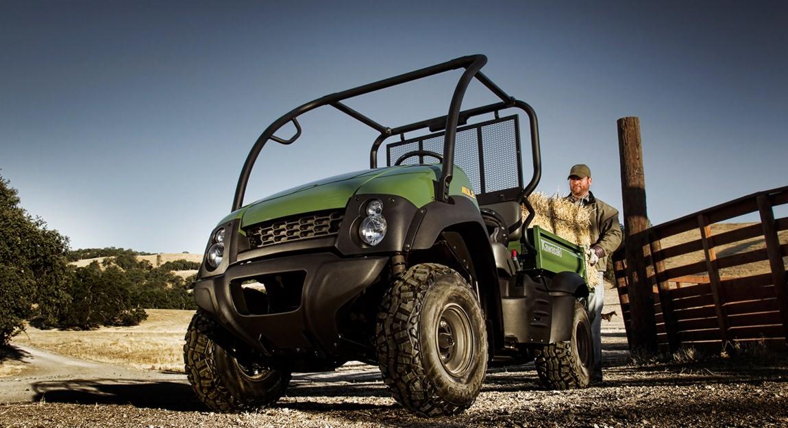 Zielony pojazd Kawasaki MULE 4010 Diesel 4x4 na polnej drodze, z mężczyzną w roboczym ubraniu i kapeluszu, trzymającym siano na pace, w tle drewniany płot i pagórkowaty krajobraz.