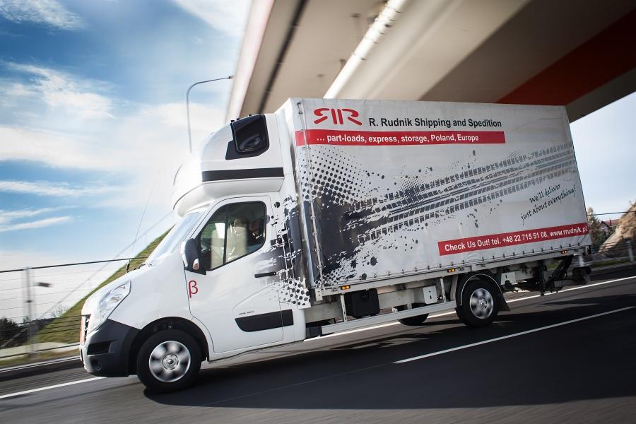 Biały bus dostawczy R. Rudnik Shipping and Spedition w ruchu, widoczny z boku na tle błękitnego nieba i konstrukcji mostu. Na naczepie grafika imitująca ślady opon.