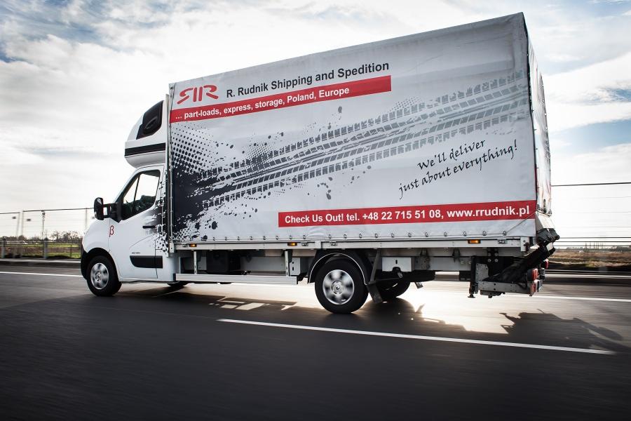 Biały bus dostawczy R. Rudnik Shipping and Spedition z grafiką imitującą ślady opon, w ruchu na asfalcie, z widocznym adresem strony internetowej.