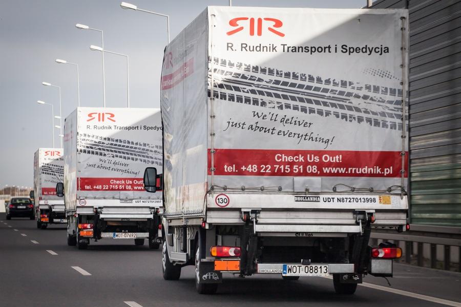 Kolumna trzech identycznych, białych busów transportowych firmy R. Rudnik Transport i Spedycja, widocznych z tyłu, jadących po autostradzie w słoneczny dzień.