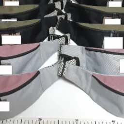 Seria różowo-szarych i czarno-zielonych saszetek biodrowych, każda z czarnym zamkiem błyskawicznym i wzorzystym paskiem, ułożonych na białym stole z podkładką do cięcia w tle.