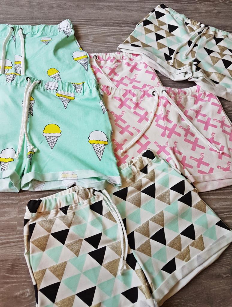 Zestaw uszytych, wzorzystych szortów dziecięcych z gumką i sznurkiem w pasie, leżących na drewnianej powierzchni. Wzory: lody, różowe krzyżyki i trójkąty w kolorach czerni, złota i mięty.