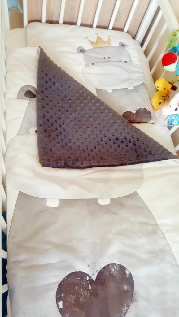 Dziecięce łóżeczko z pościelą w motywie hipopotama w koronie i szarą kołderką z wypukłymi kropkami, obok wiszą kolorowe zabawki.