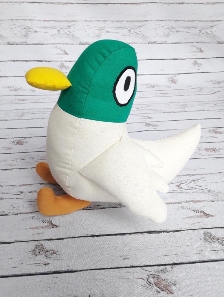 Pluszowa kaczka z zieloną głową, białym korpusem i pomarańczowymi nogami leży na drewnianej podłodze z efektem postarzenia.