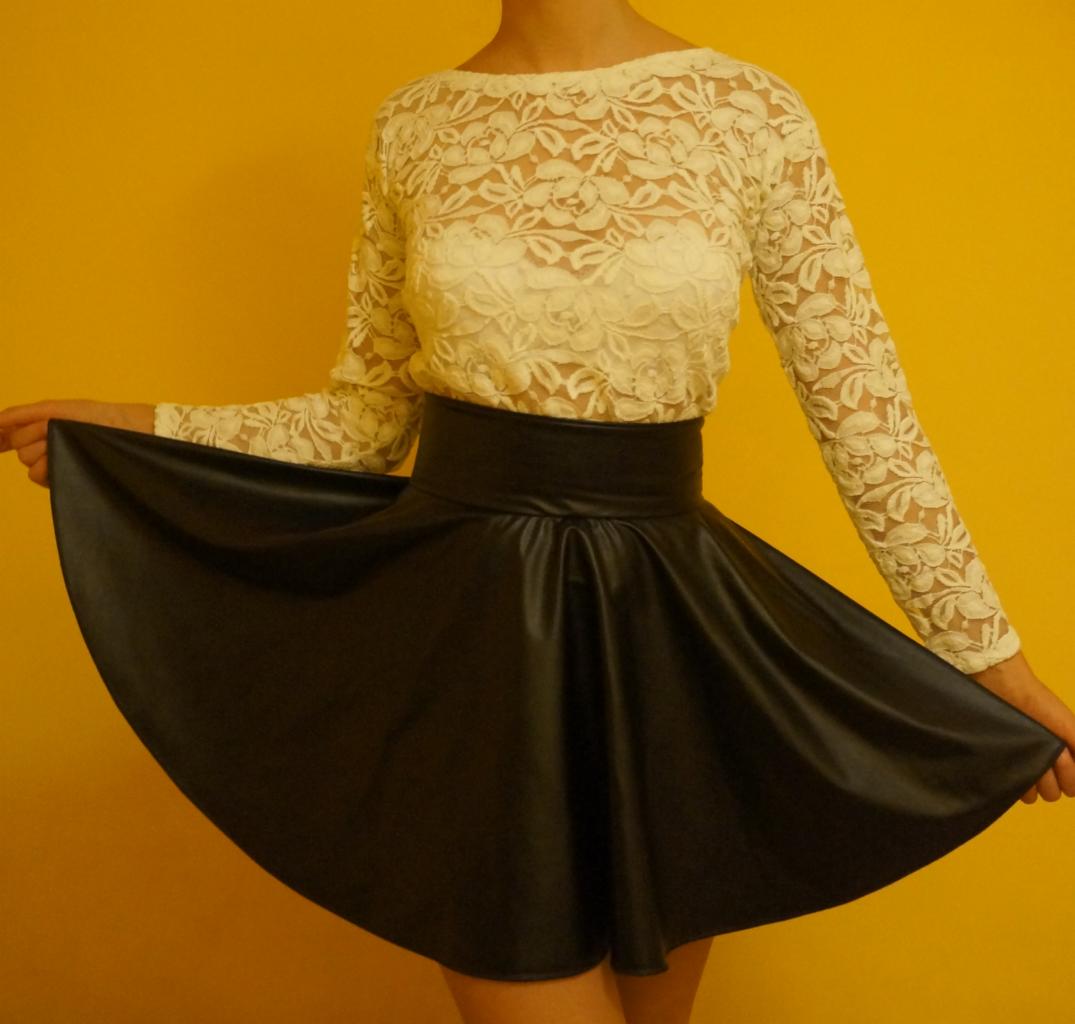 Kobieta prezentuje czarną, rozkloszowaną spódnicę z wysokim stanem i kremową bluzkę z koronki na tle żółtej ściany.