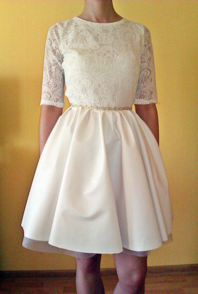 Biała sukienka z koronkową górą i rozkloszowanym dołem, przepasana delikatnym złotym paskiem, na żółtym tle.