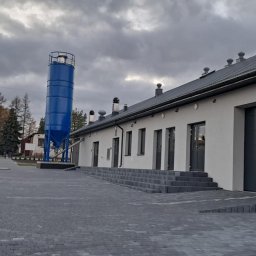 EXPRESS- BUD SPÓŁKA Z OGRANICZONĄ ODPOWIEDZIALNOŚCIĄ - Nowoczesny budynek z szarym dachem i niebieskim silosem na zewnątrz, otoczony brukowaną nawierzchnią. Widoczne schody wejściowe i brama garażowa.
