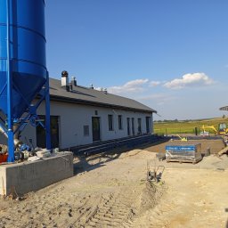 EXPRESS- BUD SPÓŁKA Z OGRANICZONĄ ODPOWIEDZIALNOŚCIĄ - Budowa hali: niebieski silos z żółtą drabinką, szara hala z kominami, przygotowany teren z kostką brukową i sprzętem budowlanym w tle, błękitne niebo.