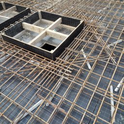 EXPRESS- BUD SPÓŁKA Z OGRANICZONĄ ODPOWIEDZIALNOŚCIĄ - Zbrojenie fundamentów z drewnianymi szalunkami, widok z góry. Siatka z prętów zbrojeniowych na czarnej folii, betonowe dystanse. Etap budowy domu.