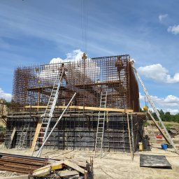 EXPRESS- BUD SPÓŁKA Z OGRANICZONĄ ODPOWIEDZIALNOŚCIĄ - Zbrojenie fundamentów pod budynek w trakcie realizacji. Widoczne szalunki, stalowe pręty, drabiny i pracownicy na rusztowaniu. Praca wre w słoneczny dzień.