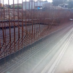 EXPRESS- BUD SPÓŁKA Z OGRANICZONĄ ODPOWIEDZIALNOŚCIĄ - Skomplikowana konstrukcja zbrojeniowa z prętów stalowych, przygotowana do zalania betonem. W tle widoczny ciężki sprzęt budowlany i materiały budowlane.