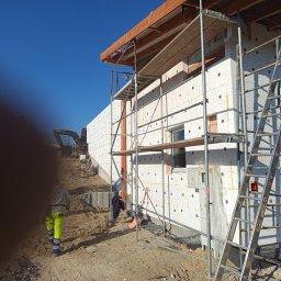 EXPRESS- BUD SPÓŁKA Z OGRANICZONĄ ODPOWIEDZIALNOŚCIĄ - Budowa domu: ściany ocieplone styropianem, rusztowania, drabina. W tle koparka i pracownik w odblaskowych spodniach. Etap ocieplania elewacji w słoneczny dzień.