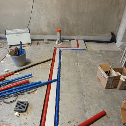 EXPRESS- BUD SPÓŁKA Z OGRANICZONĄ ODPOWIEDZIALNOŚCIĄ - Montaż instalacji ogrzewania podłogowego: czerwone i niebieskie rury w wyżłobieniach, szary beton, grzejnik, wiadro z narzędziami, kartony. Etap prac przed wylaniem posadzki.
