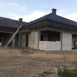 Budowa domu z białych bloczków, widoczne rusztowania i drabina. Częściowo położony dach z czarnej dachówki. Dom w stanie surowym otwartym, piasek na podwórku.