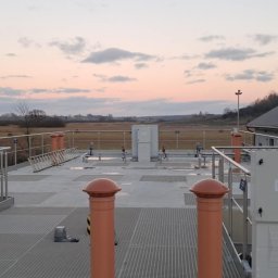 Instalacja przemysłowa na dachu budynku z widokiem na rozległe tereny pod zachmurzonym niebem. Widoczne rury, zawory, skrzynki elektryczne i drabina.