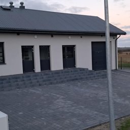 Nowoczesny, parterowy budynek z szarym dachem i kostką brukową. Widoczne trzy wejścia z drzwiami w kolorze grafitowym. W tle pole z belami siana i zachodzące słońce.