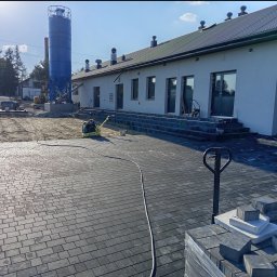 EXPRESS- BUD SPÓŁKA Z OGRANICZONĄ ODPOWIEDZIALNOŚCIĄ - Układanie kostki brukowej na posesji w Krasnobród. Widoczna ubijarka, paleta z kostką i budynek w tle. Kostka ułożona do połowy powierzchni.