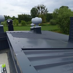 EXPRESS- BUD SPÓŁKA Z OGRANICZONĄ ODPOWIEDZIALNOŚCIĄ - Szary dach z blachy trapezowej z kominkami wentylacyjnymi na tle zieleni. Widoczne mocowania blachy. Dach po montażu, gotowy do użytku.