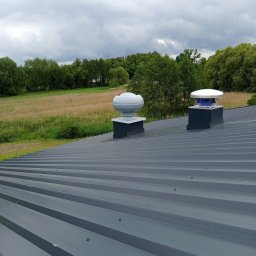 EXPRESS- BUD SPÓŁKA Z OGRANICZONĄ ODPOWIEDZIALNOŚCIĄ - Szary dach z blachy trapezowej z dwoma kominkami wentylacyjnymi na tle zieleni i pochmurnego nieba. Widok z góry, równomierne oświetlenie.