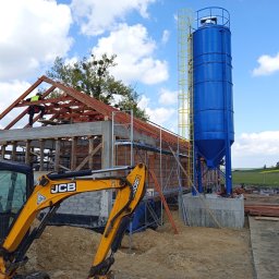 EXPRESS- BUD SPÓŁKA Z OGRANICZONĄ ODPOWIEDZIALNOŚCIĄ - Budowa domu z cegły: szkielet dachu w trakcie montażu, niebieski silos na zaprawę i żółta koparka JCB na pierwszym planie. Widoczne rusztowania i elementy konstrukcyjne.