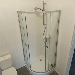 EXPRESS- BUD SPÓŁKA Z OGRANICZONĄ ODPOWIEDZIALNOŚCIĄ - Narożna kabina prysznicowa z chromowanymi profilami i szklanymi drzwiami, zamontowana w łazience z białymi płytkami i szarą podłogą. Widoczny fragment toalety.